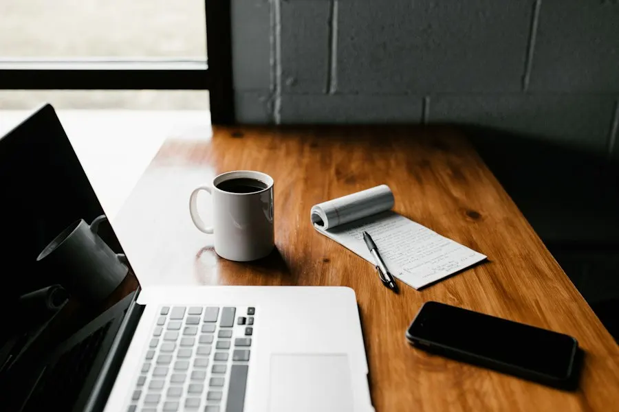 Laptop, coffee mug, phone and a notepad of handwritten notes on a wooden desk
