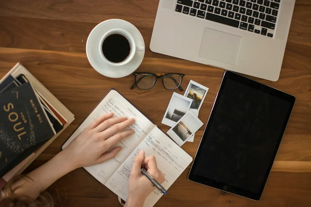 Overhead view of a desk with a laptop, a tablet, a cup of coffee and someone writing in a notebook